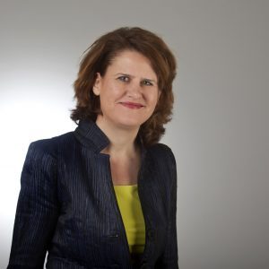 Portretfoto van een persoon in donkere blazer, lichtgele top en met middellang haar, voorzitter van een organisatie.