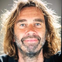 Portret van een volwassene met lang haar en baard, die glimlacht tegen een witte achtergrond met lichtsporen.