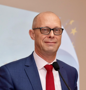 Voorzitter Cees van Dam spreekt met microfoon, in donkerblauwe jas, wit overhemd en rode das tegen een achtergrond met logo.