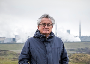 Een oudere persoon met bril en donkerblauwe jas staat voor een industriële installatie met schoorstenen.